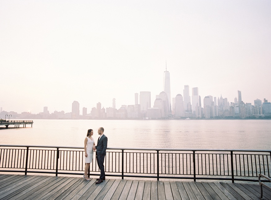 Elina and Thomas walking hand in hand along the Exchange Place waterfront at sunrise.