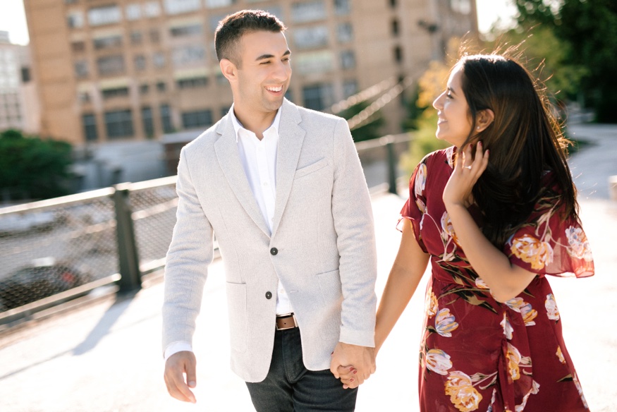 Roma and Eric strolling along the white stone pathway at The Rail Park, city buildings on both sides.