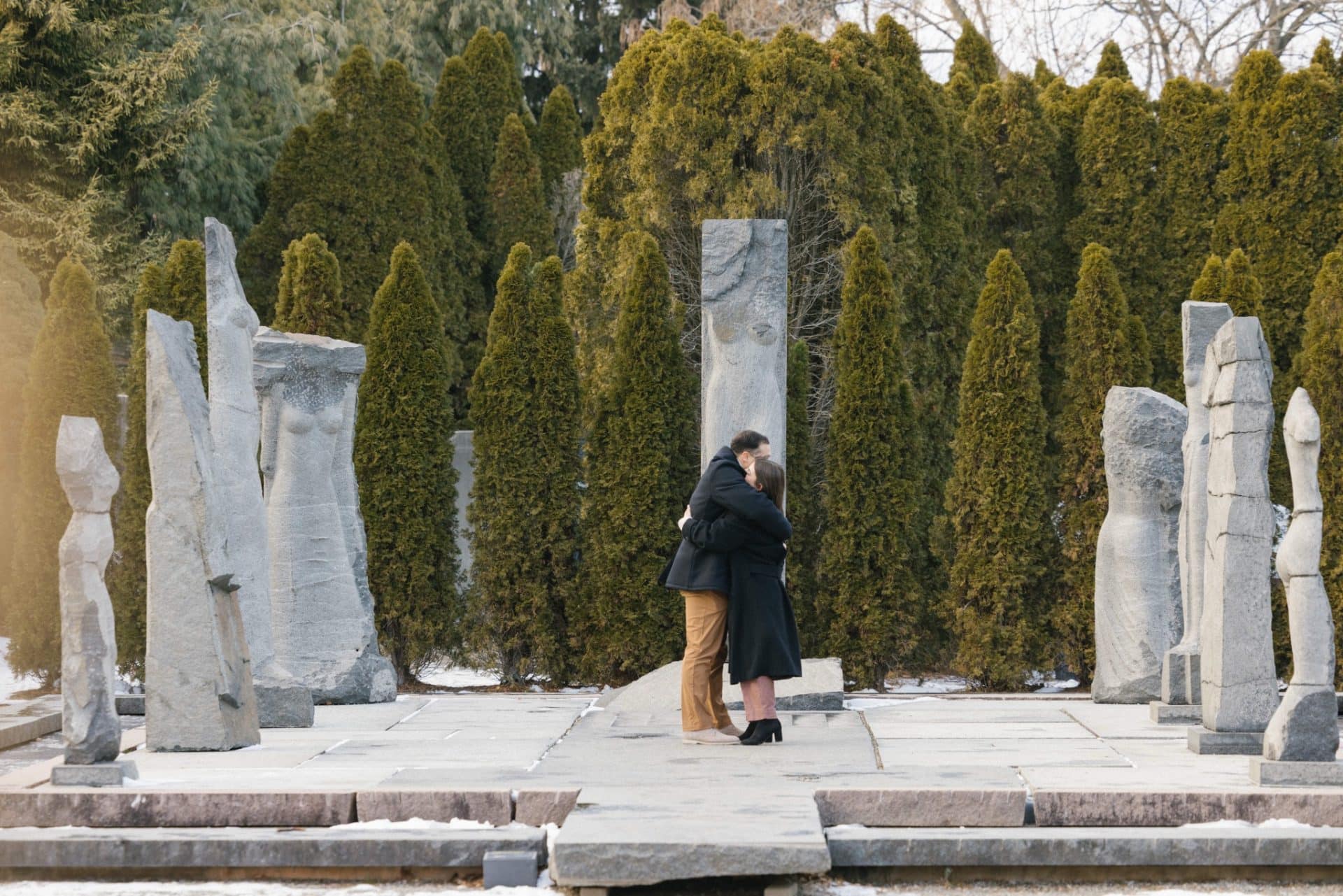 Couple embrace among sculptures after winter proposal at Grounds for Sculpture Nine Muses Hamilton NJ - snow proposal photographer