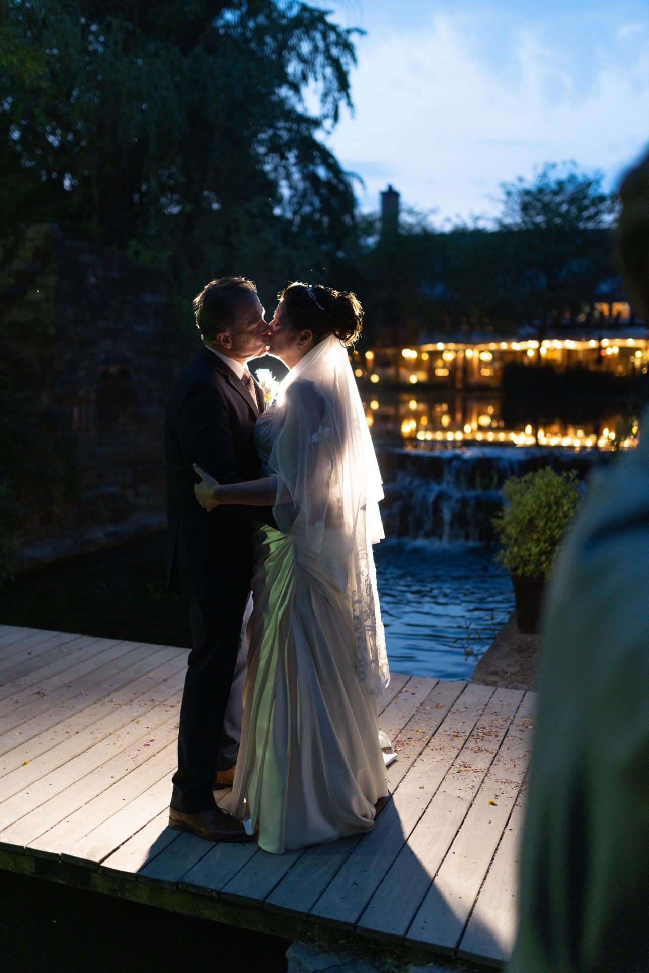 Couple kissing on the bridge at night with Rat's Restaurant lit up and waterfall reflection at Grounds for Sculpture in Hamilton NJ