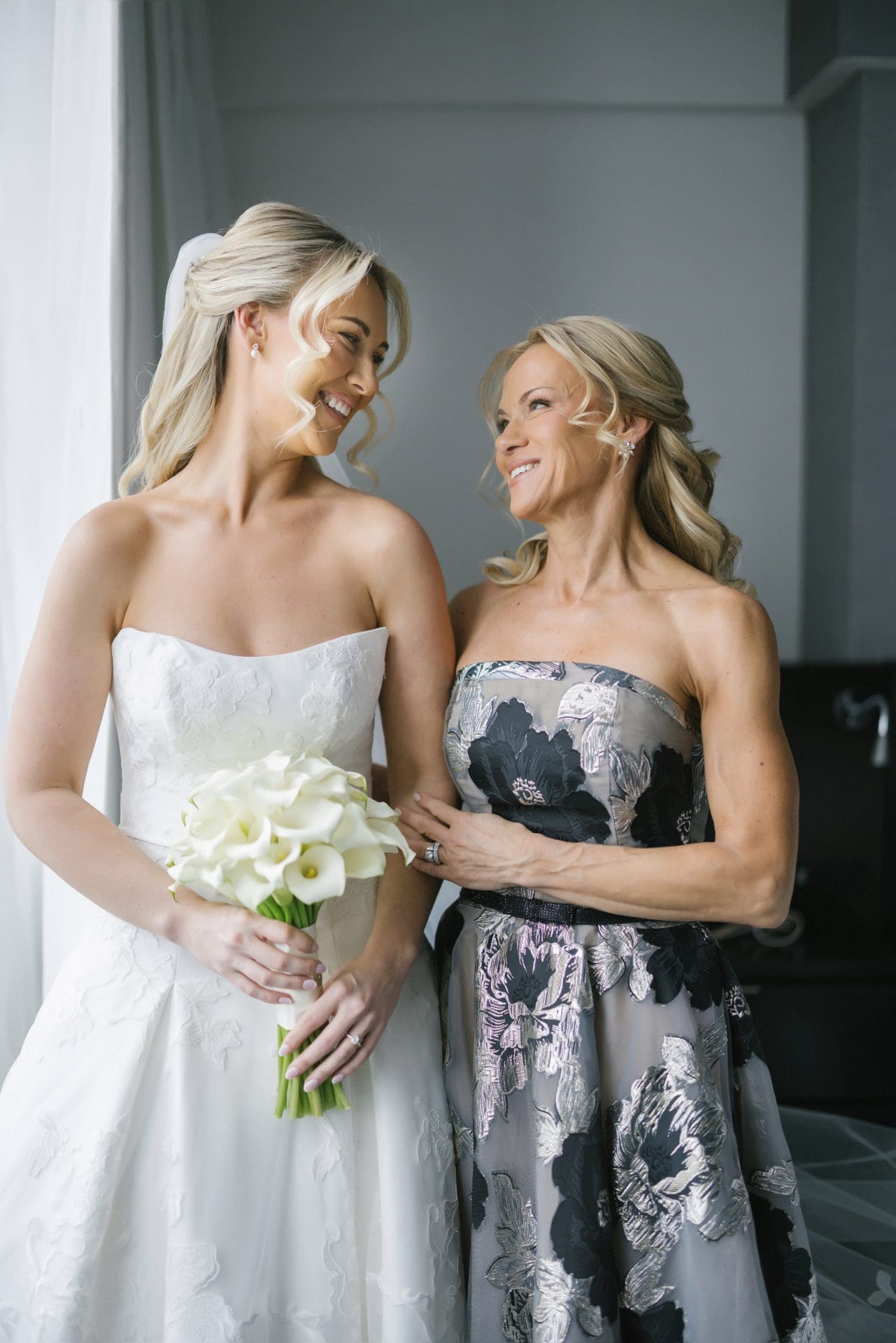 Bride and mother smiling at each other holding white calla lily bouquet