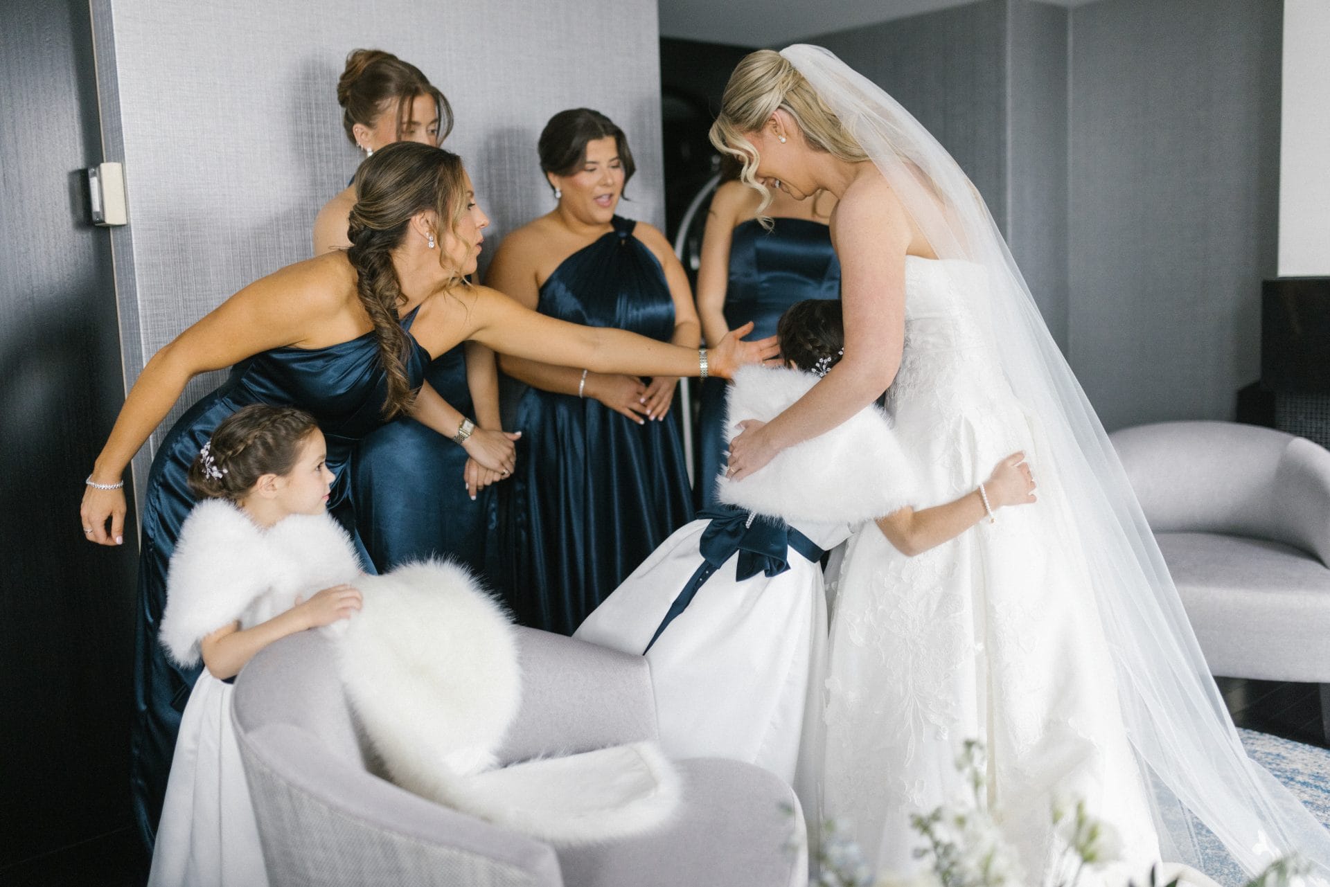 Bride reaching toward flower girl while bridesmaids look on in hotel suite