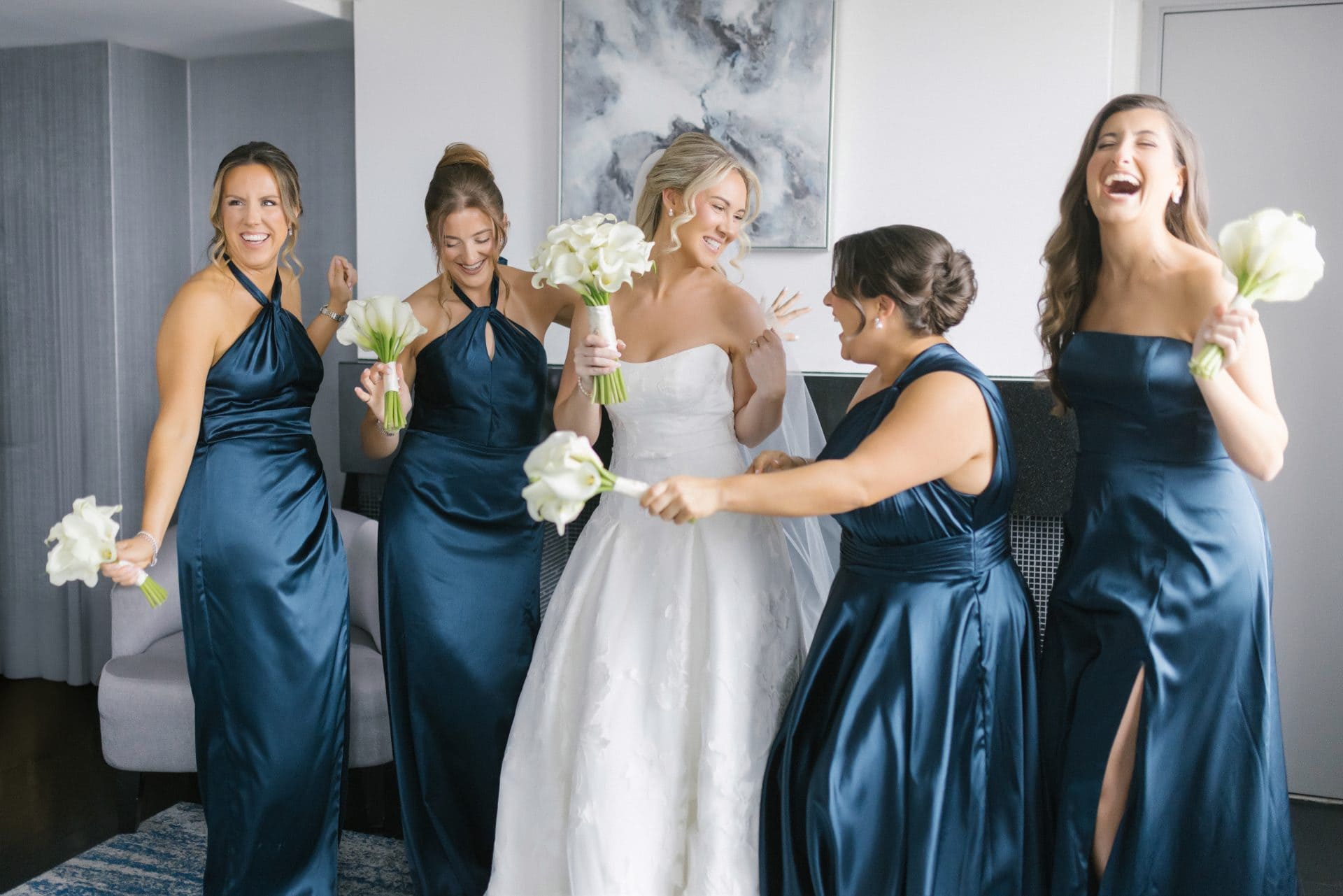 Bridal party in navy satin gowns laughing together holding white calla lily bouquets