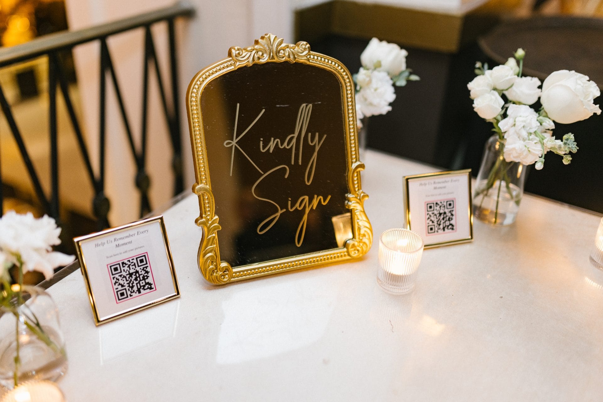 Kindly sign gold mirror with QR codes and white flowers at wedding reception