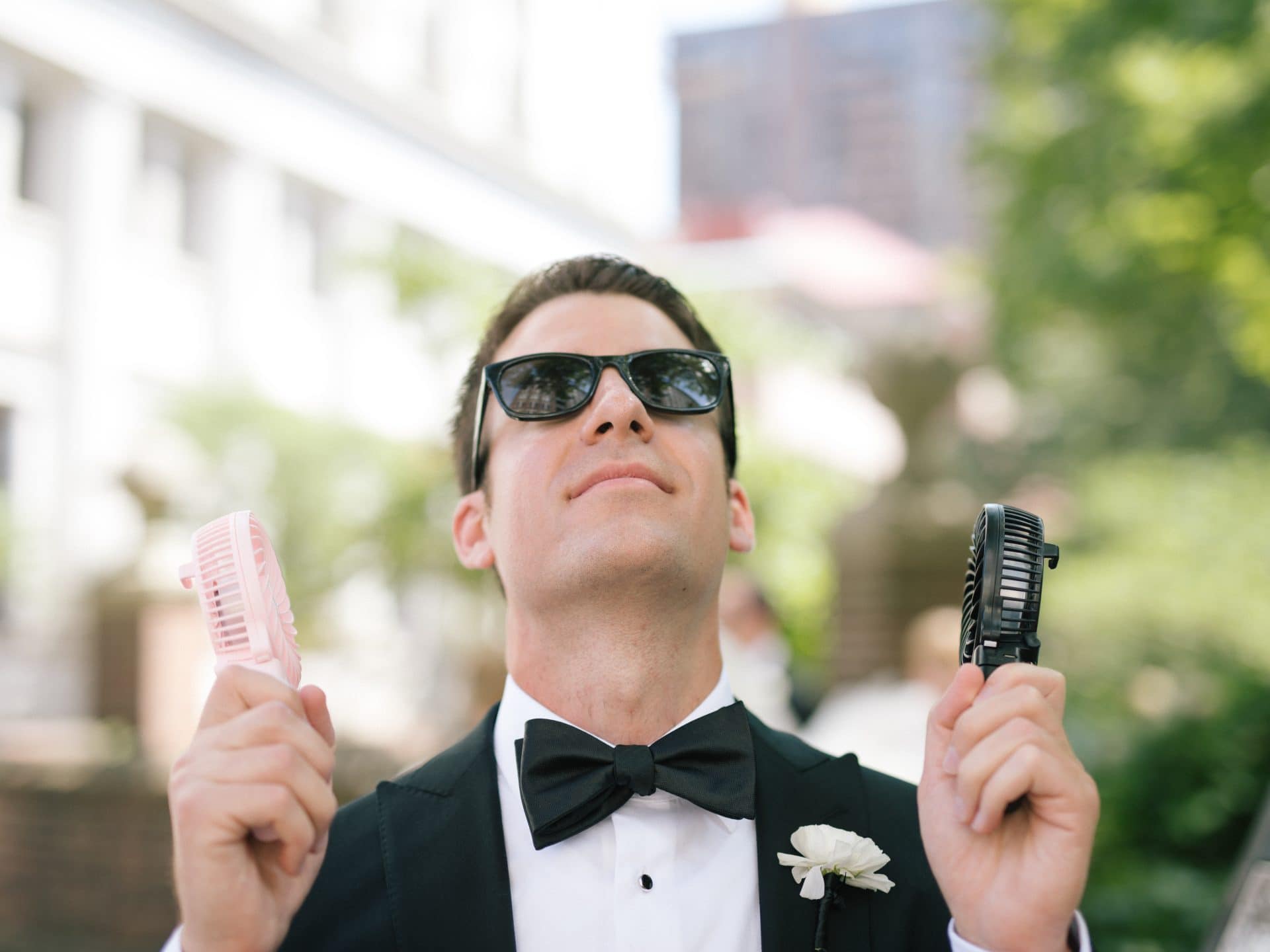 Groom in a tuxedo and sunglasses grinning with a handheld fan in each hand on a hot wedding day