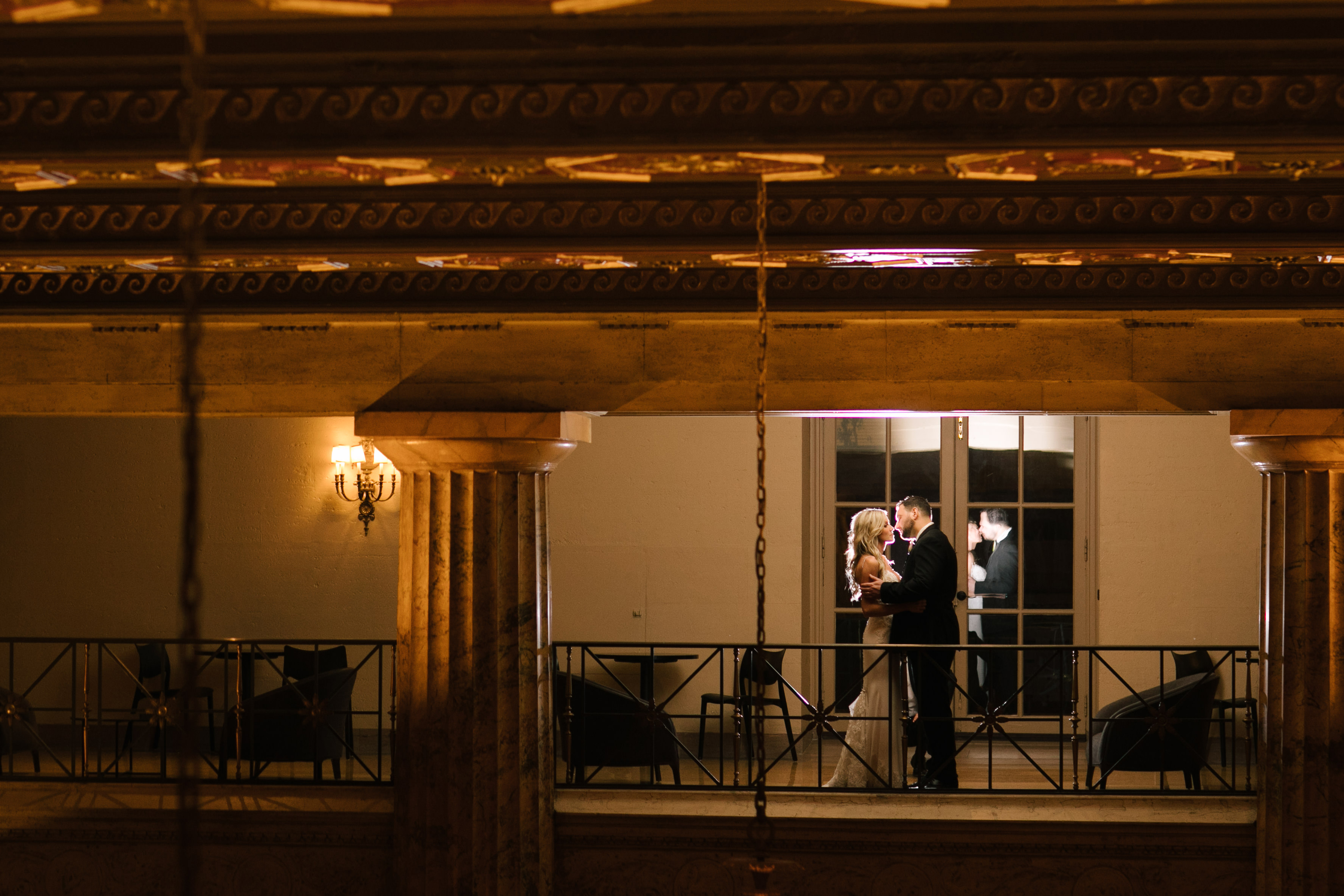 Bride and groom holding each other on the lobby mezzanine balcony at the Ballroom at the Ben in Philadelphia, framed through the columns and brass railing in warm evening light, photographed by Jordan Brian