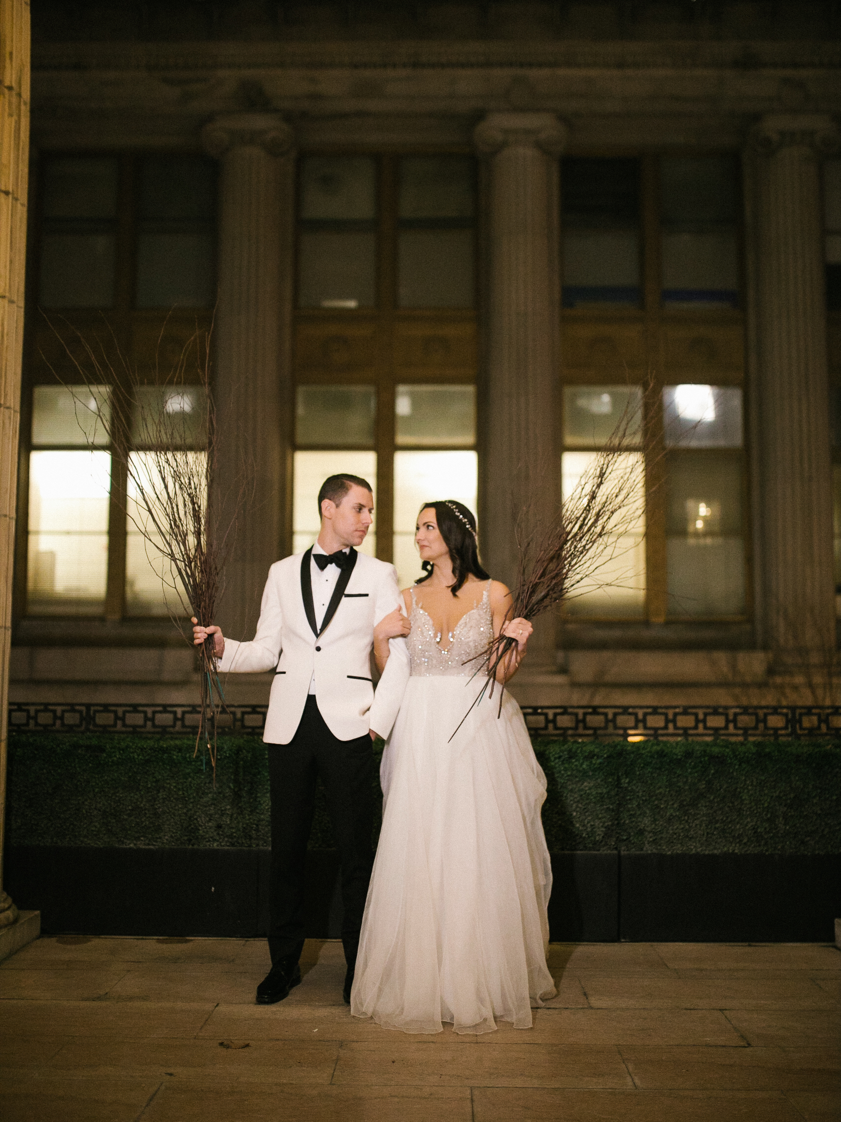 Laura and Michael on the Ballroom at the Ben balcony at night, each holding a bundle of bare branches, photographed by Jordan Brian