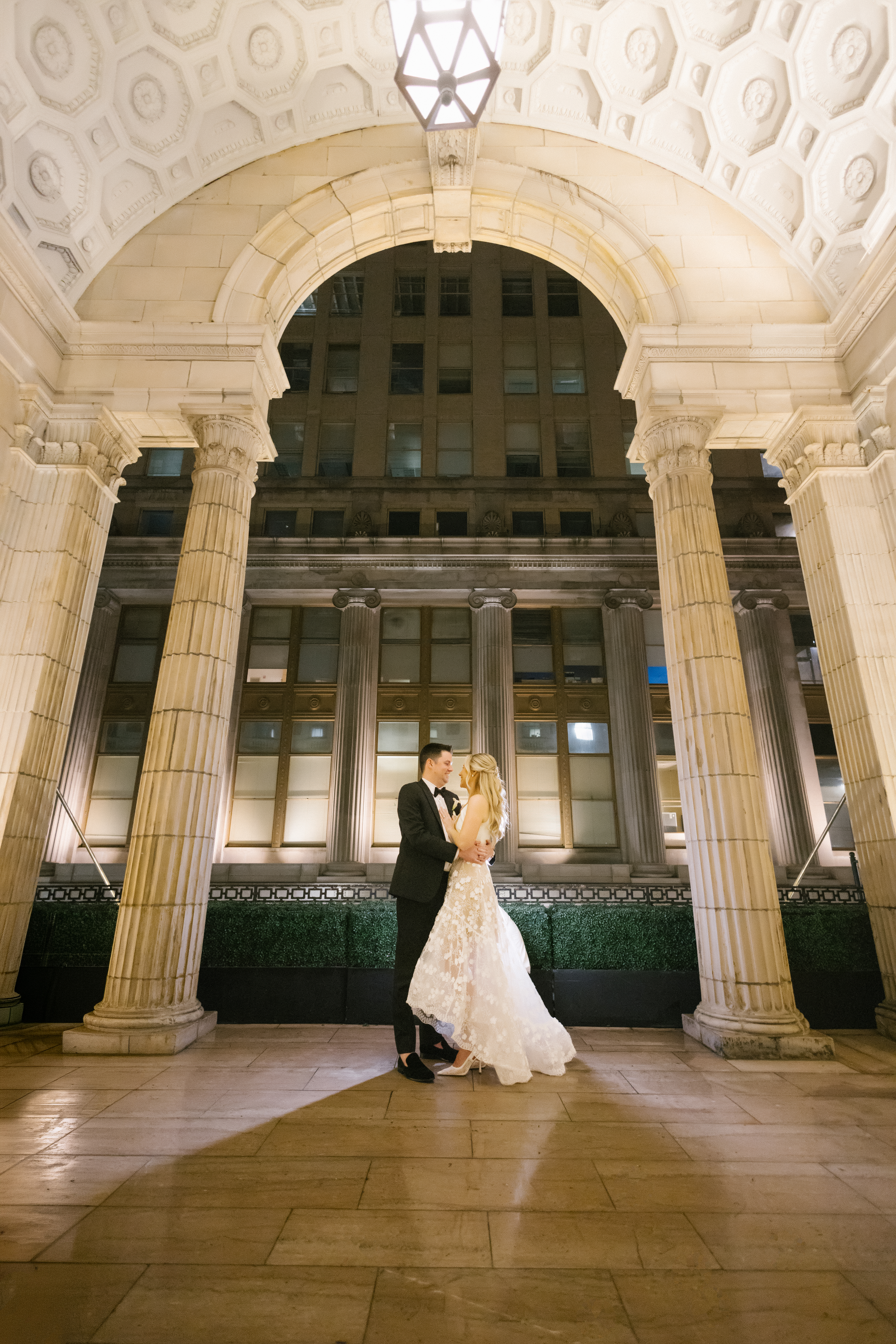 Bride and groom under the lit stone arch on the outdoor balcony at the Ballroom at the Ben in Philadelphia at night, columns framing the city behind them, photographed by Jordan Brian