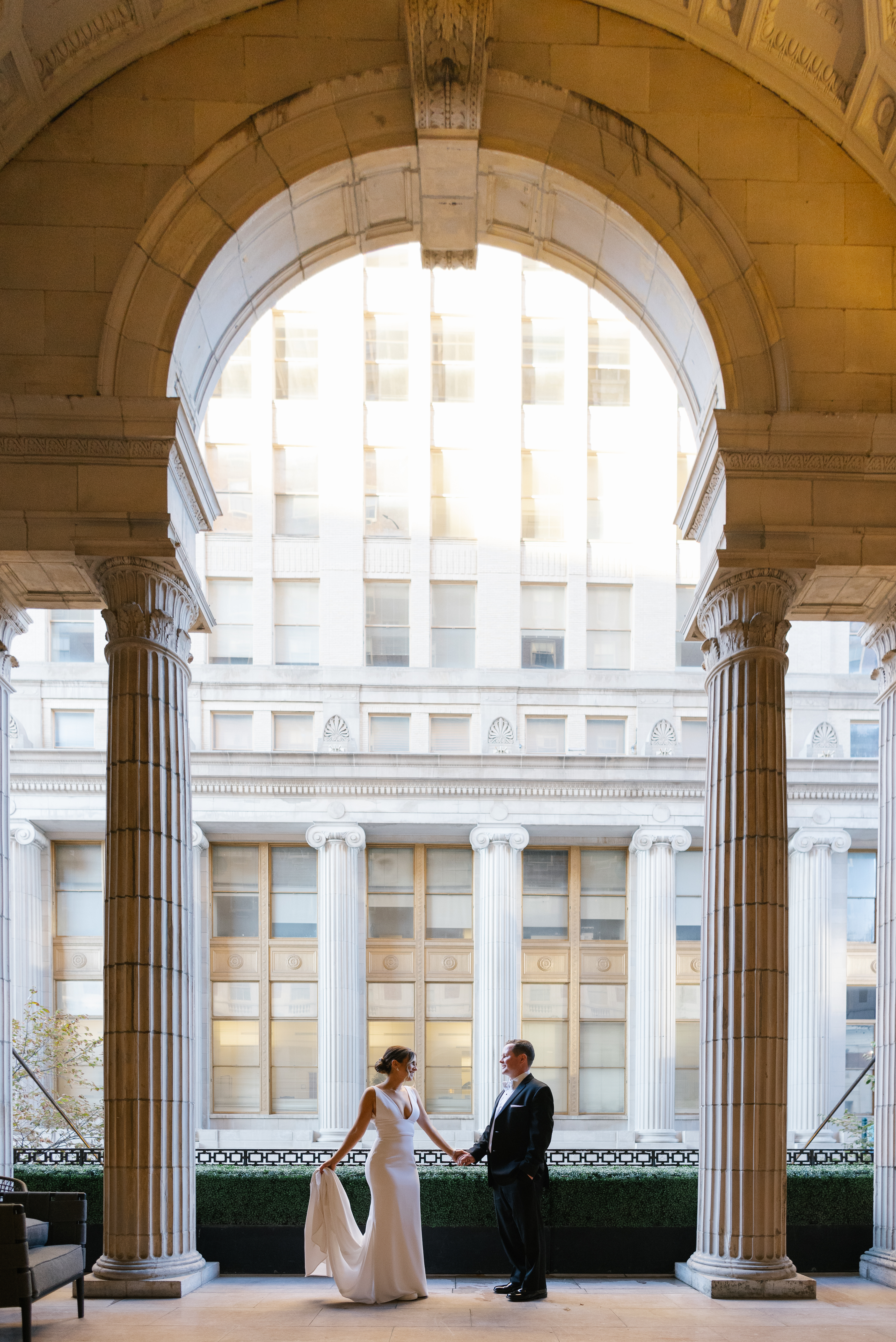 Bride and groom holding hands under the same stone arch on the outdoor balcony at the Ballroom at the Ben in Philadelphia in daylight, Center City buildings bright behind them, photographed by Jordan Brian
