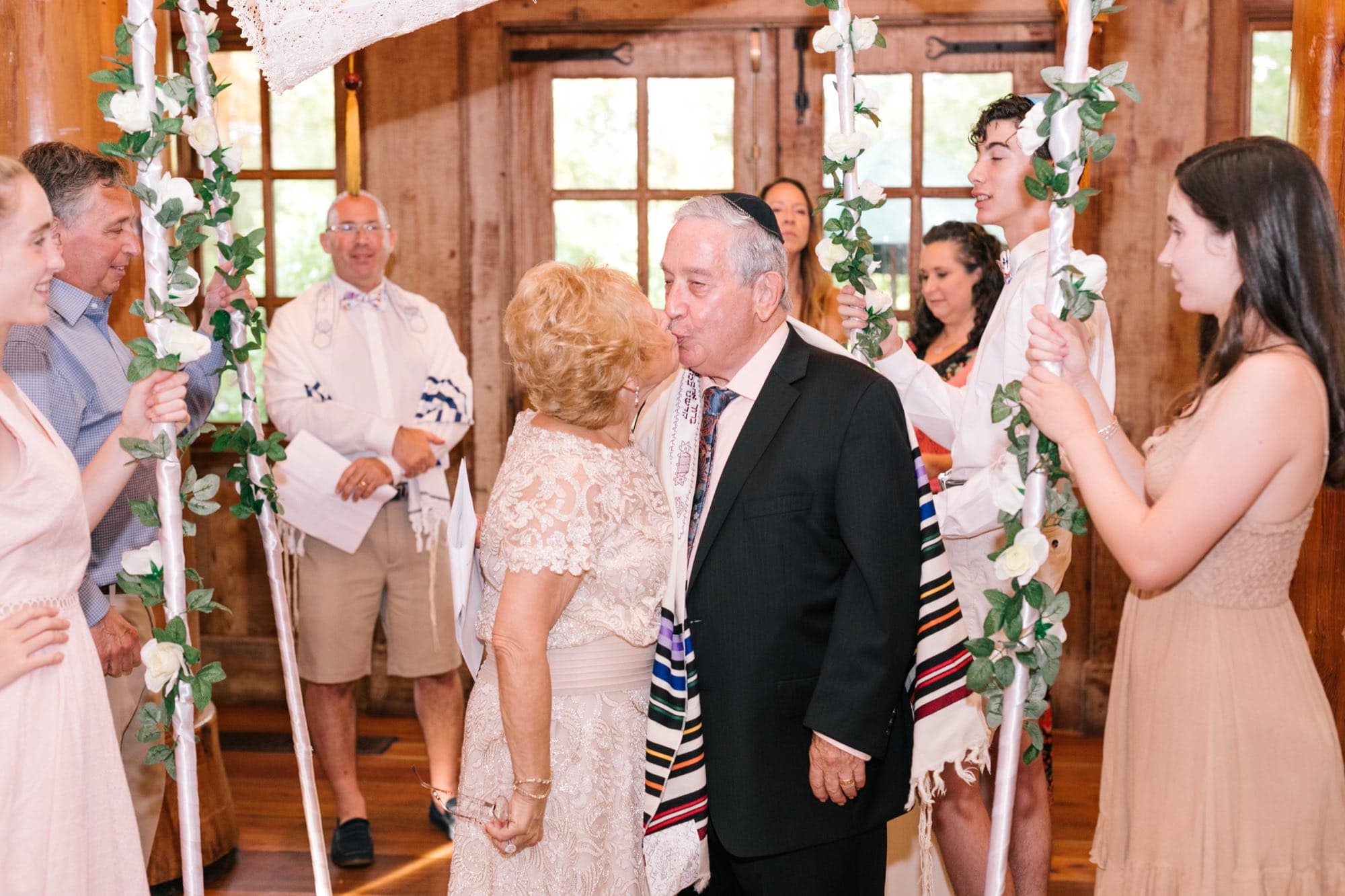 An older couple kissing under a tallit held as a chuppah during their fiftieth anniversary vow renewal