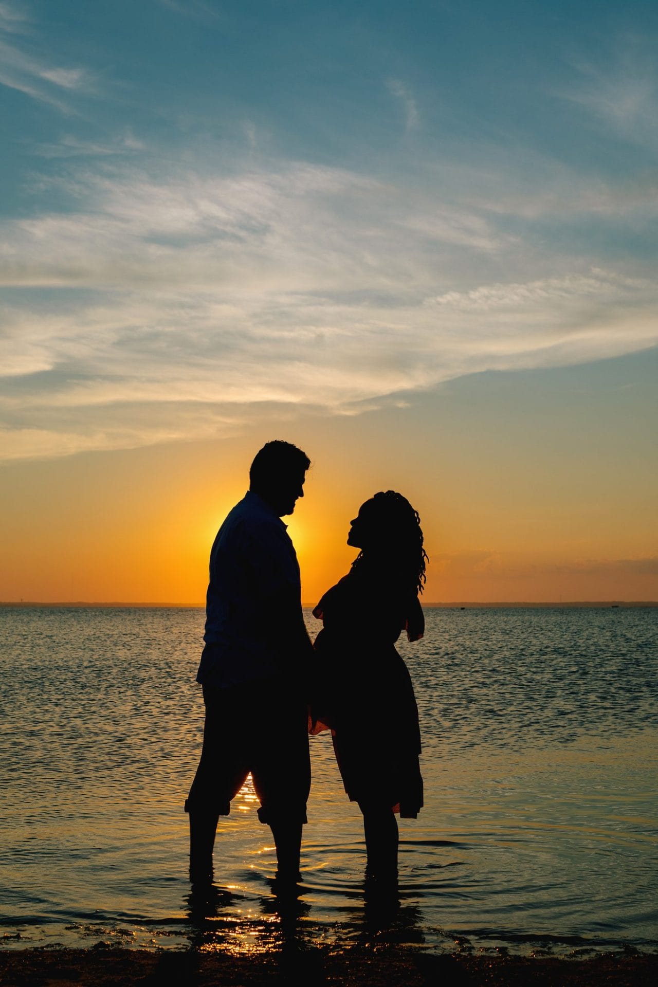 A husband and wife facing each other in silhouette with the sun between them over the bay