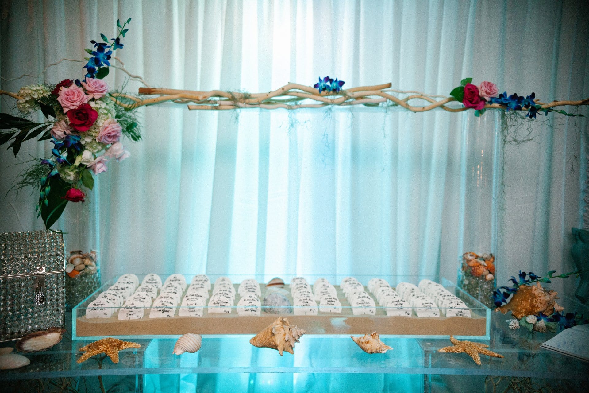 Escort cards hand lettered on seashells, set in sand beneath a driftwood and orchid arch