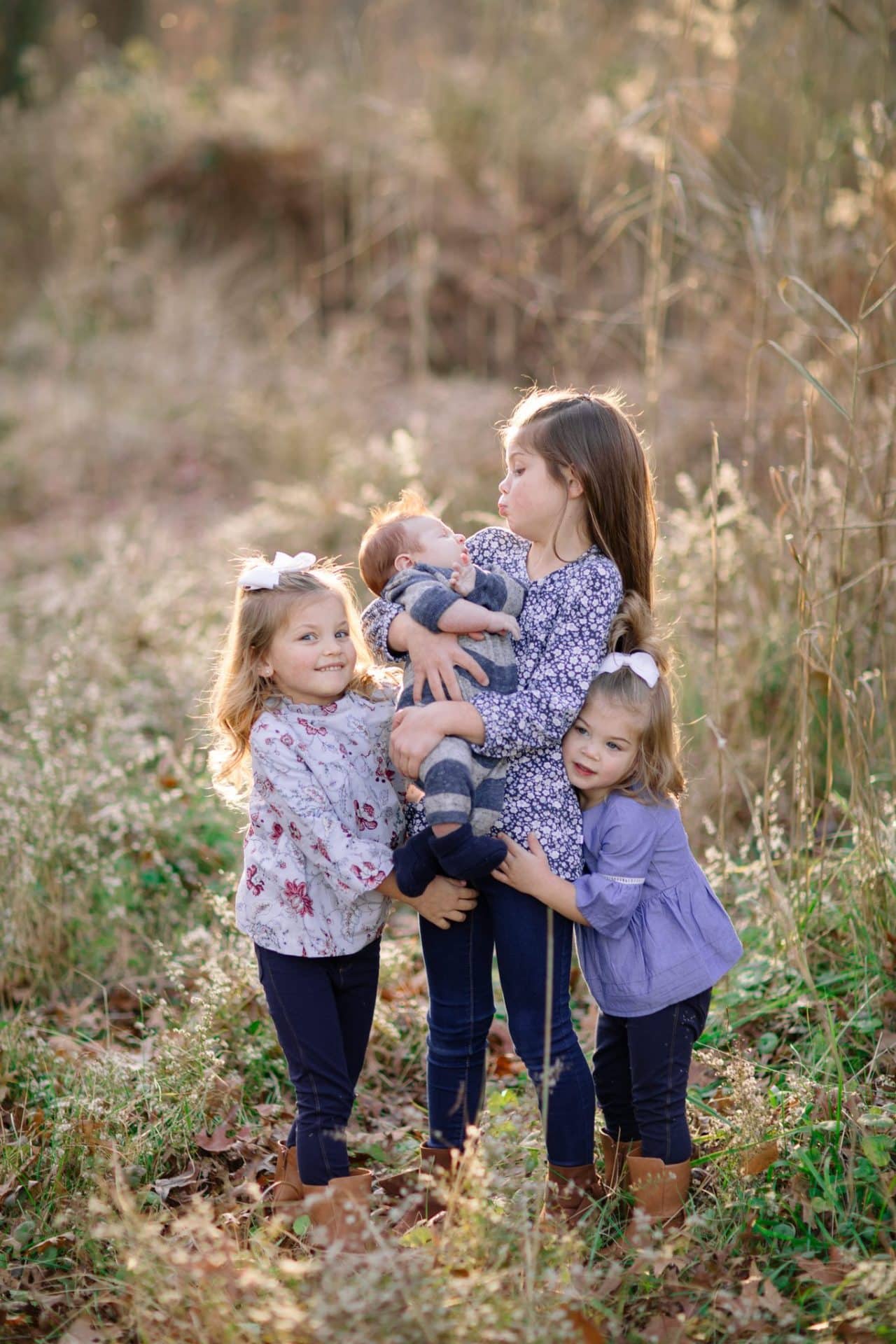 Three sisters in the tall grass with the baby, the oldest leaning down to kiss the top of the baby's head, in Medford, New Jersey