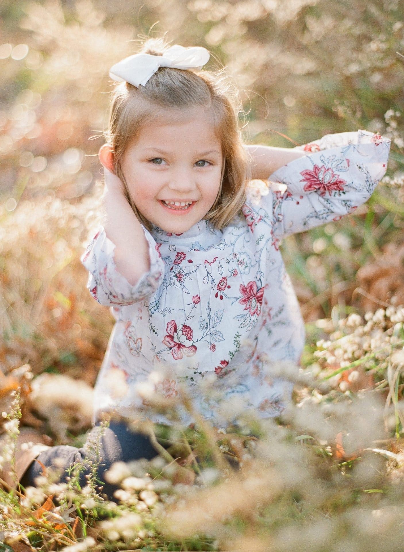 A young girl sitting low in tall backlit seed grass with both hands behind her head in Medford, New Jersey