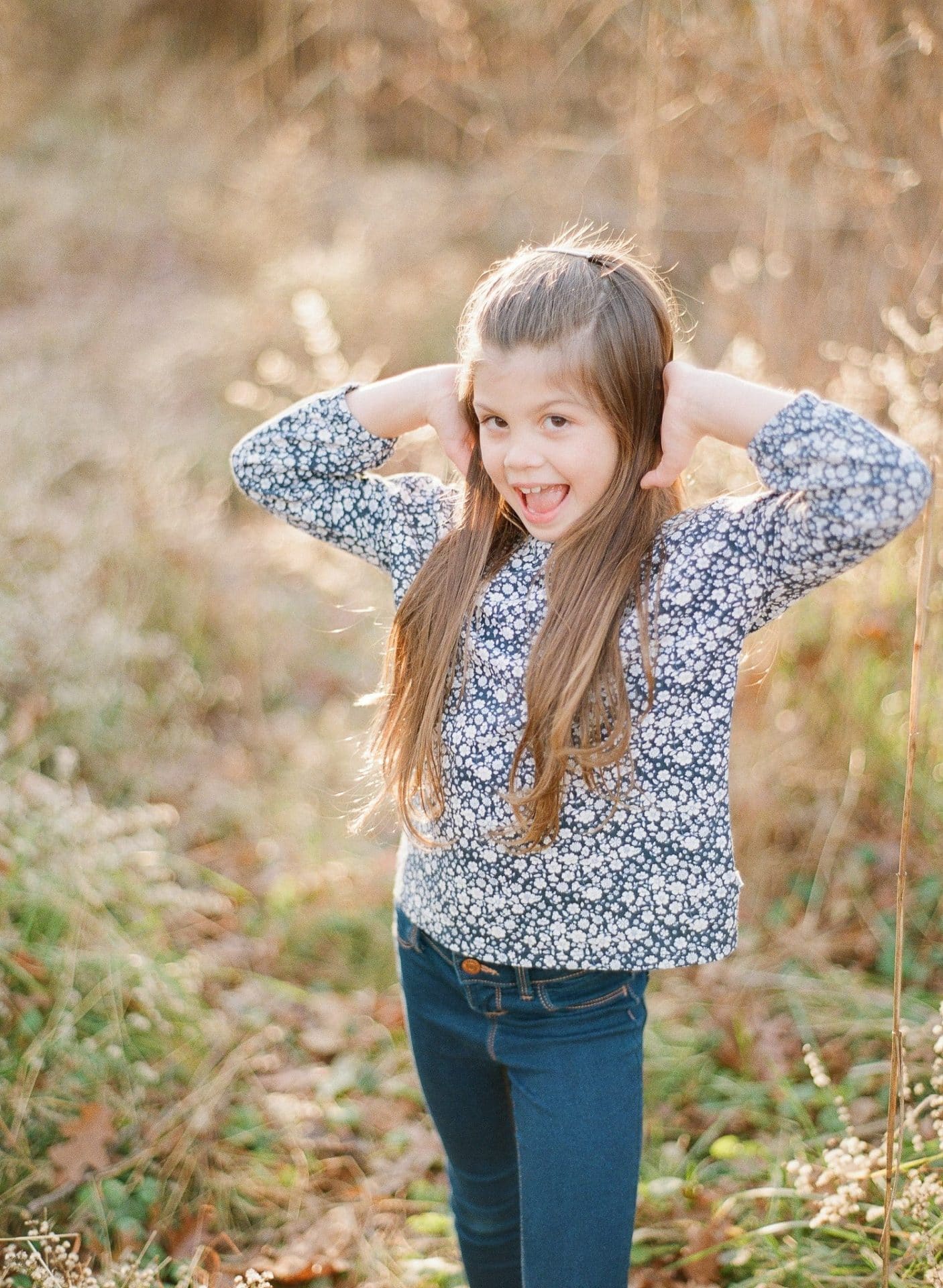 An older sister laughing with both hands up behind her ears in a field of tall backlit grass in Medford, New Jersey