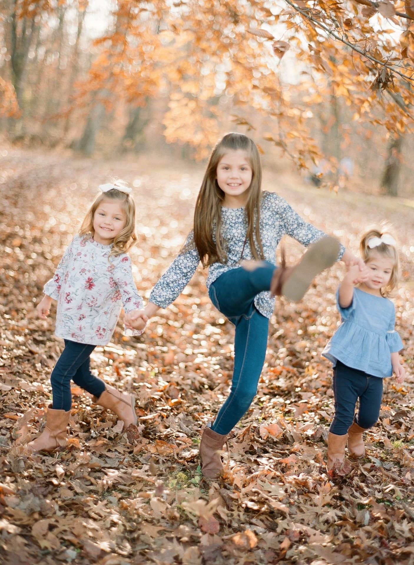 Three sisters standing in ankle deep leaves, the oldest kicking a bootful of them into the air, in Medford, New Jersey