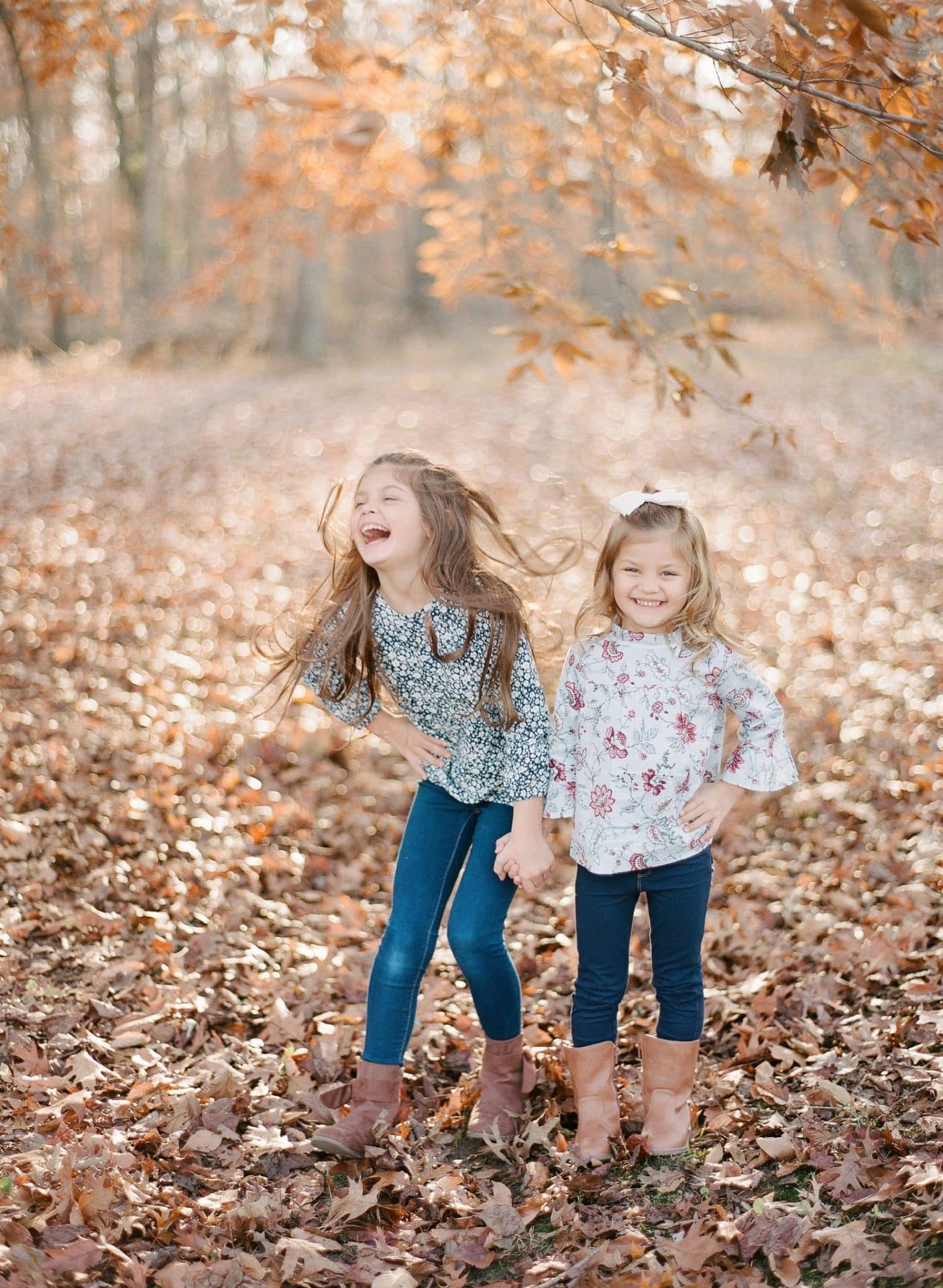 Two older sisters holding hands in the leaves, one of them laughing with her head back, during a fall session in Medford, New Jersey