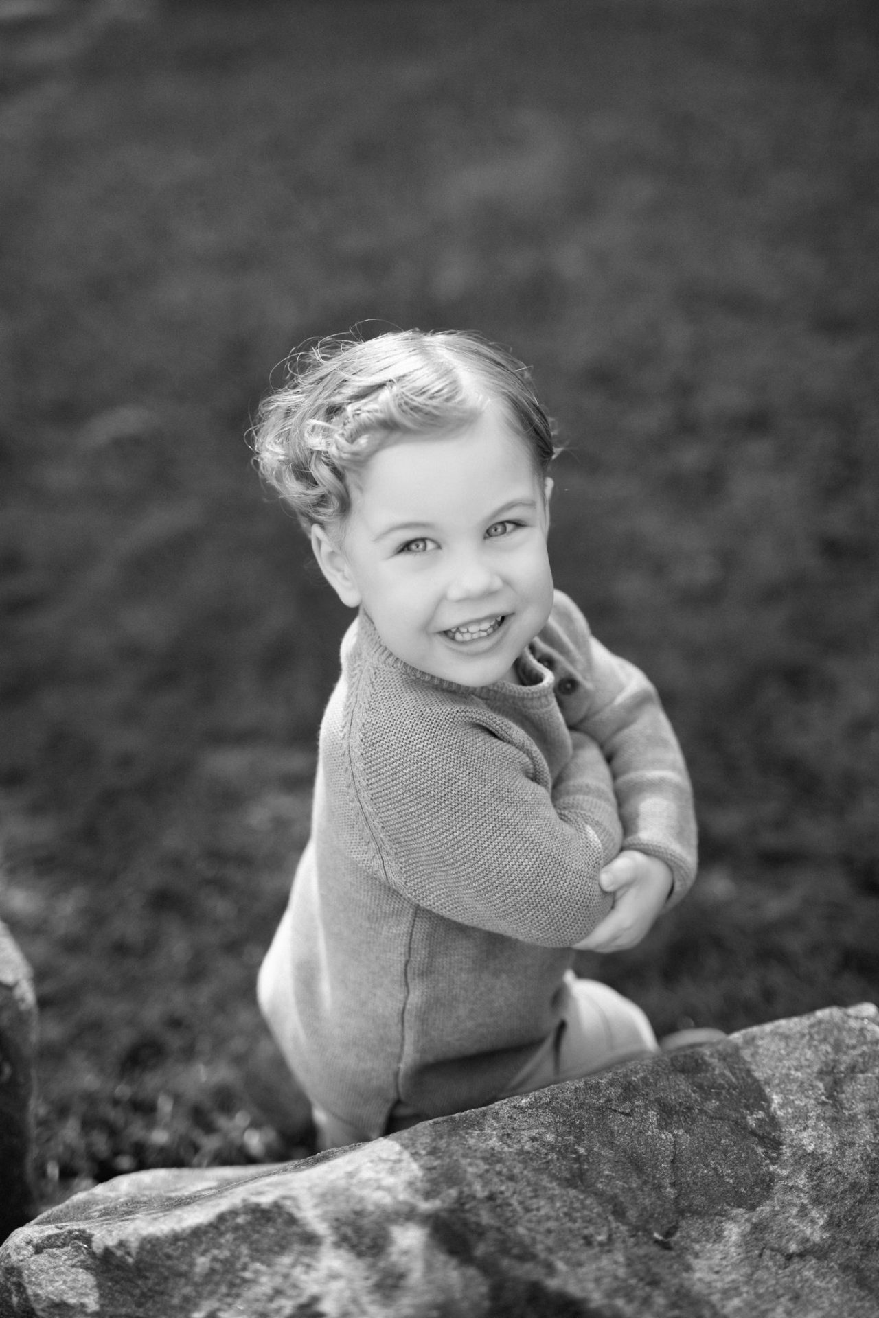 A black and white photograph of a small boy kneeling in the grass behind a rock with his arms folded, looking back over his shoulder, in Mount Laurel, New Jersey