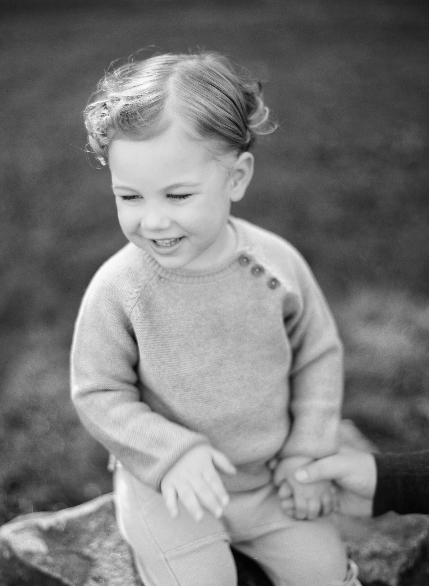 A black and white photograph of a small boy sitting on a rock and laughing, with a parent holding his hand at the edge of the frame, in Mount Laurel, New Jersey