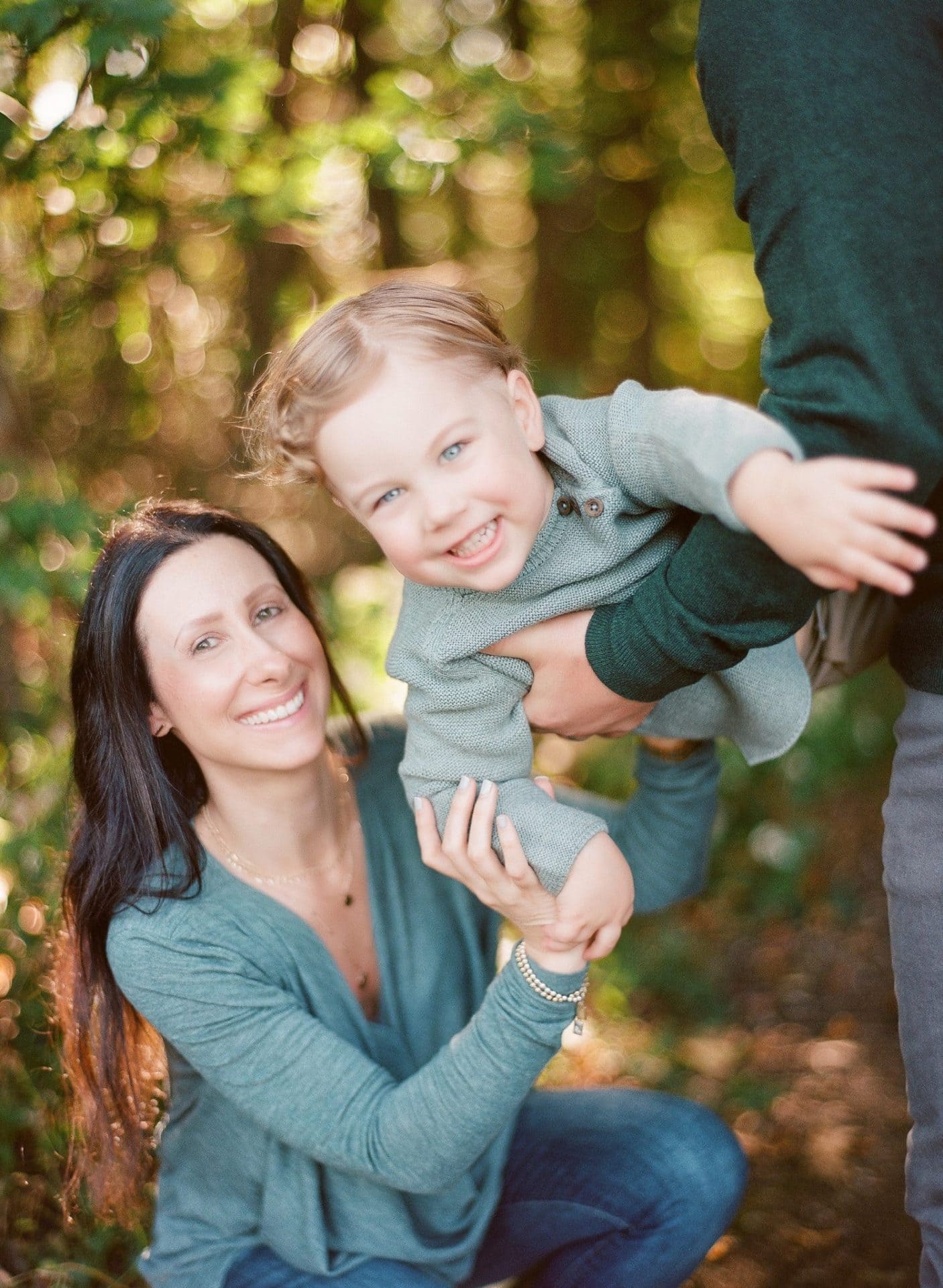 A mom holding her laughing son out horizontally in front of her, his dad's hand steadying him at the edge of the frame, during a family session in Mount Laurel, New Jersey