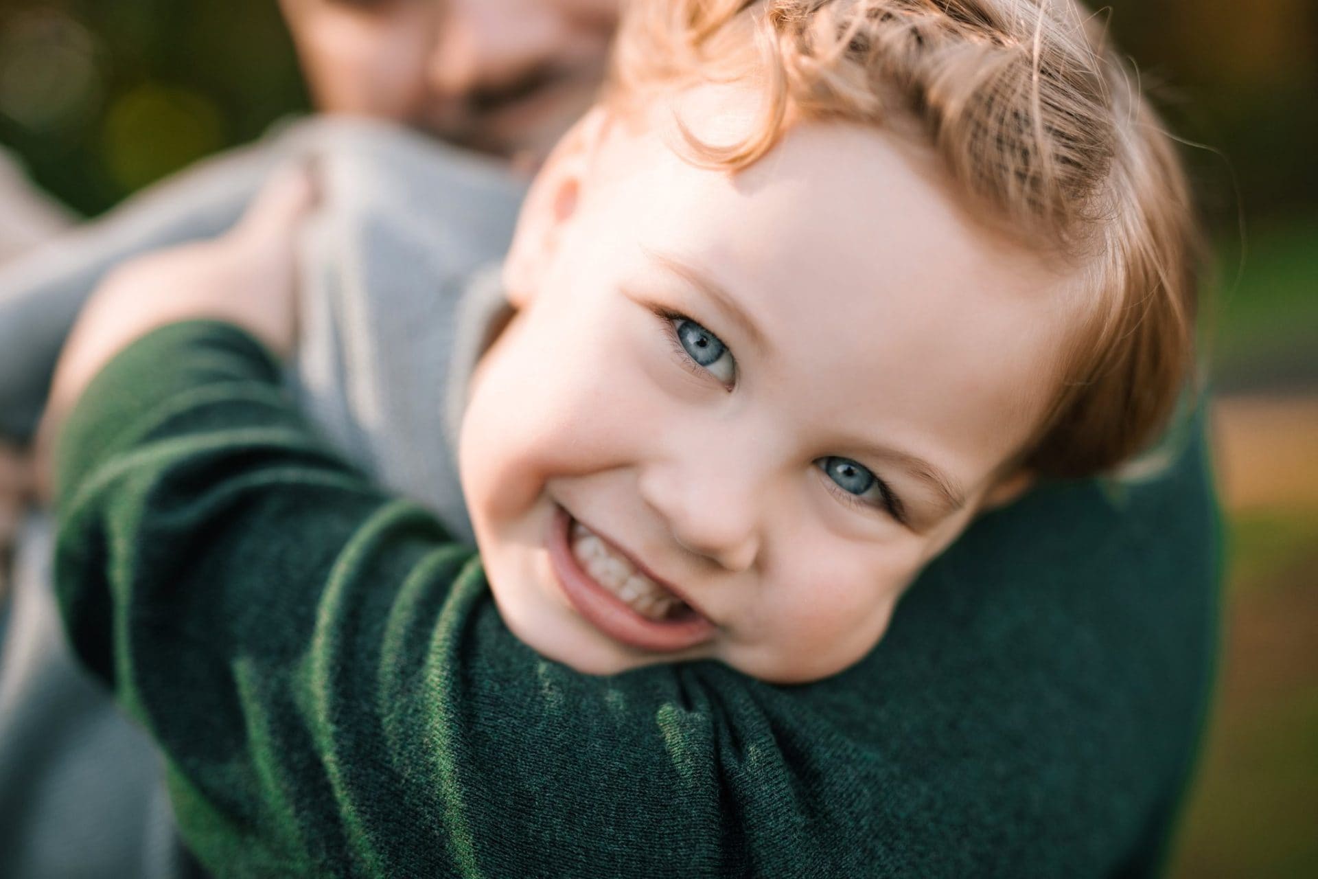 A small boy grinning over his dads shoulder during a family portrait session in Mount Laurel, New Jersey