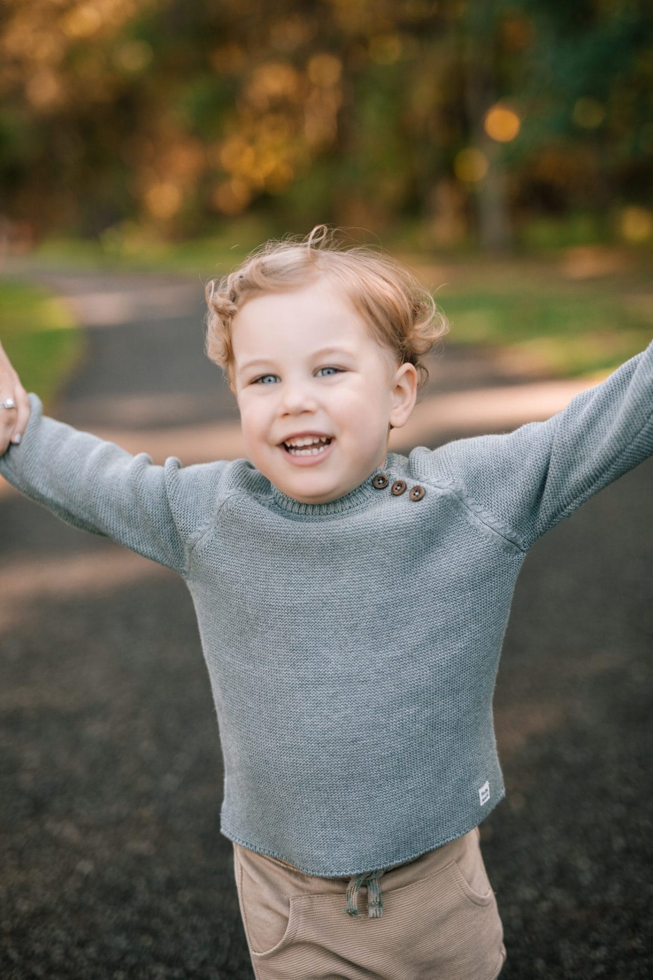 A small boy on the paved path with both arms stretched out wide, his mom's hand still holding his at the edge of the frame, at Laurel Acres Park in Mount Laurel, New Jersey