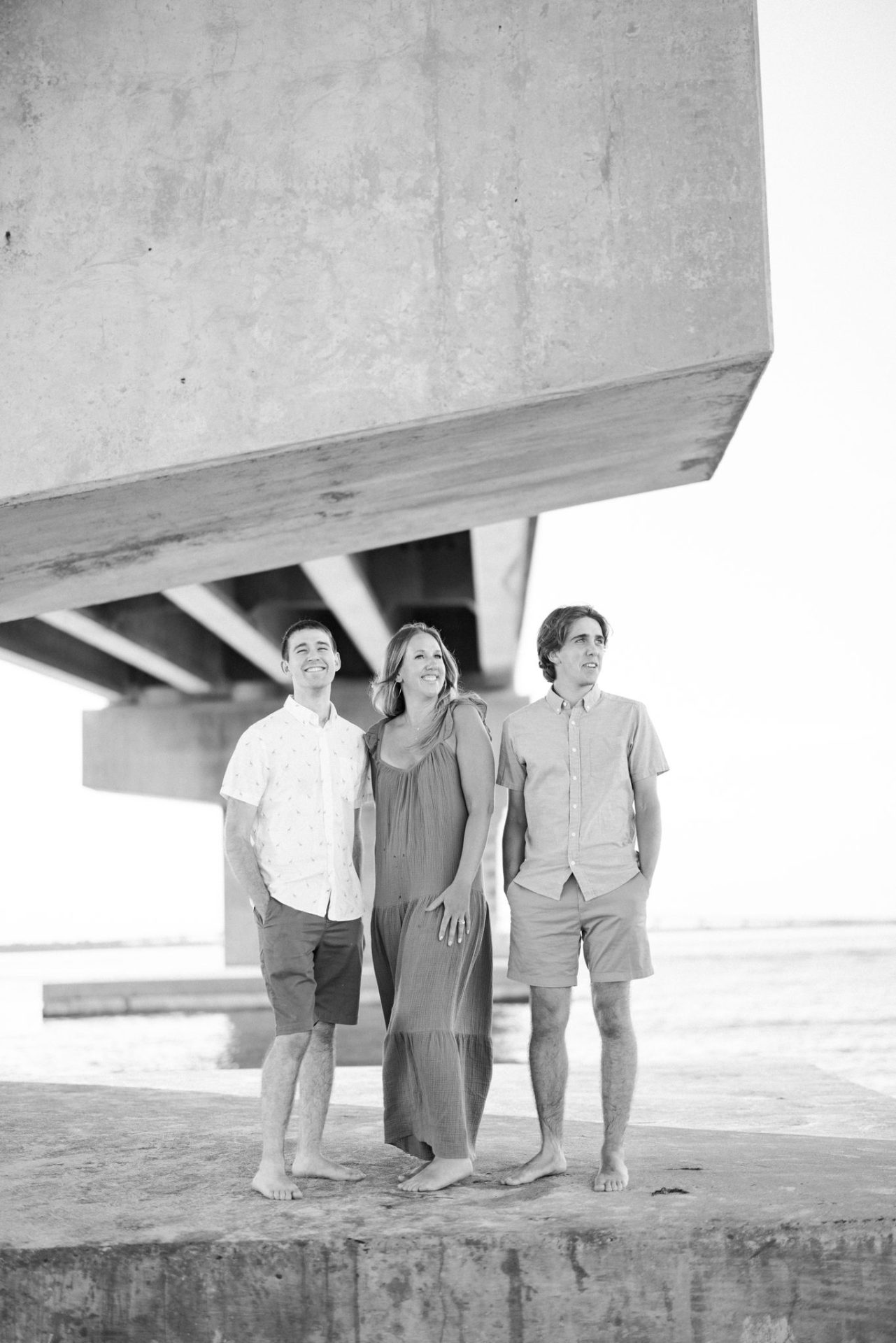 A black and white photograph of three family members standing barefoot on a concrete abutment with the underside of the bridge filling the frame above them in Ocean City, New Jersey
