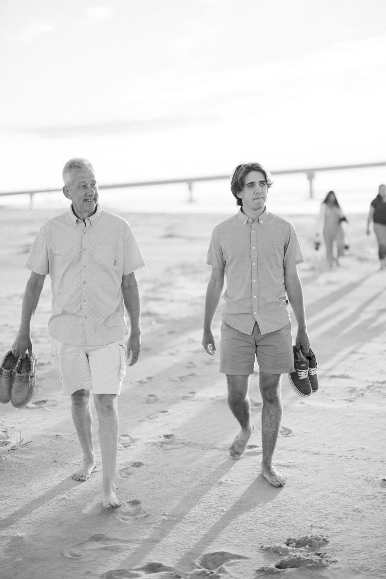 A black and white photograph of two family members walking barefoot with their shoes in hand, the bridge behind them in Ocean City, New Jersey