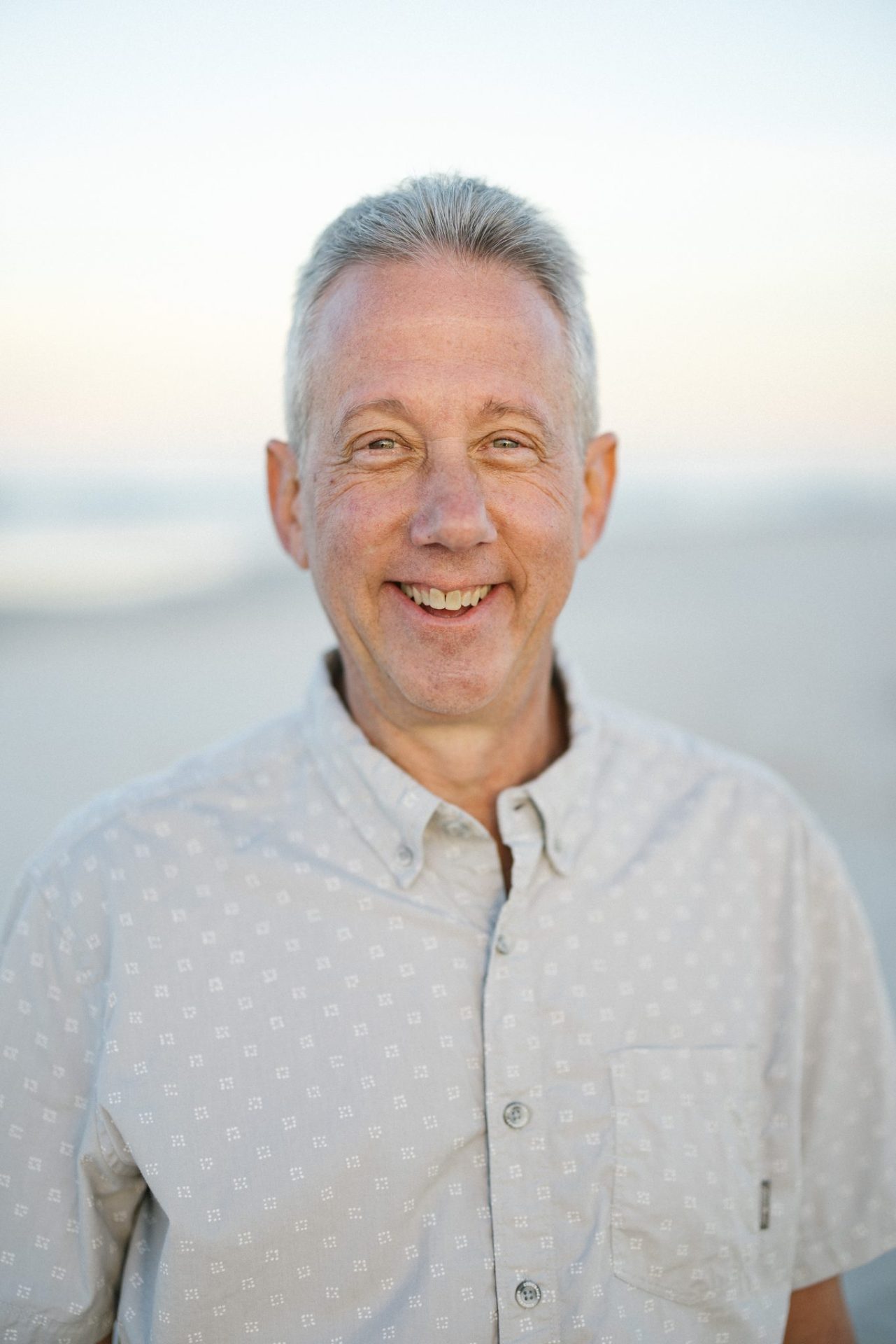 An individual portrait of a laughing man in a patterned cream button down shirt with the ocean soft behind him in Ocean City, New Jersey