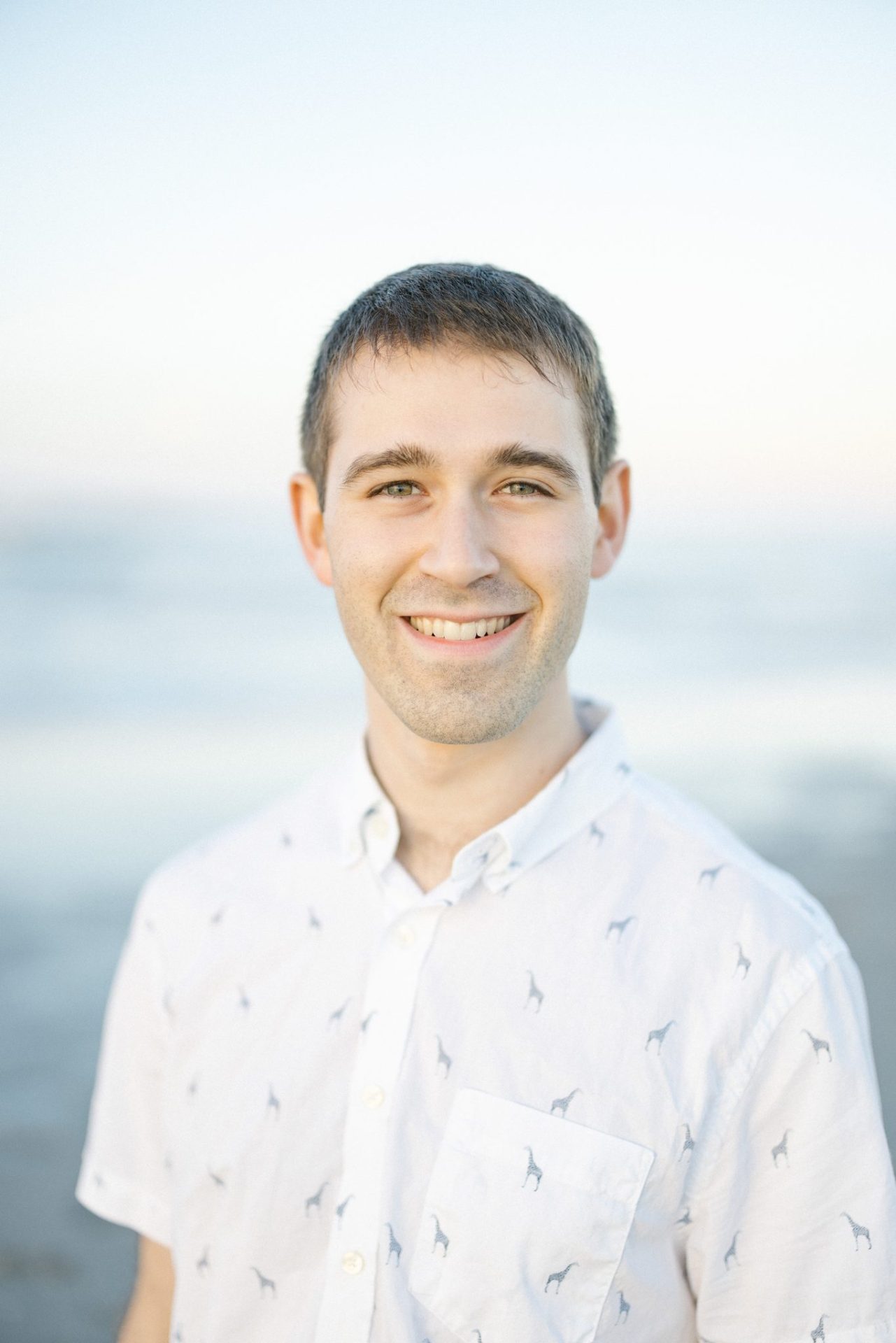 An individual portrait of a young man in a white printed button down shirt with the water soft behind him in Ocean City, New Jersey
