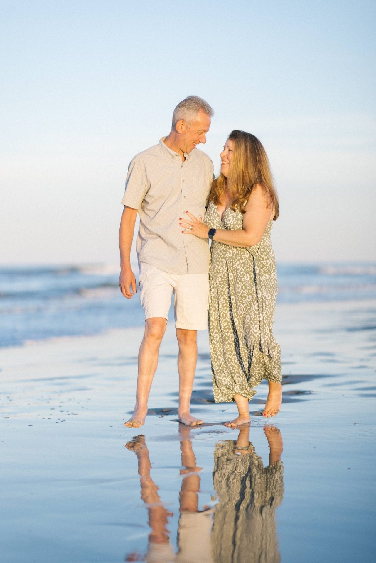 A mom and dad walking barefoot through the shallow water with her arm around him and their reflection underneath them in Ocean City, New Jersey