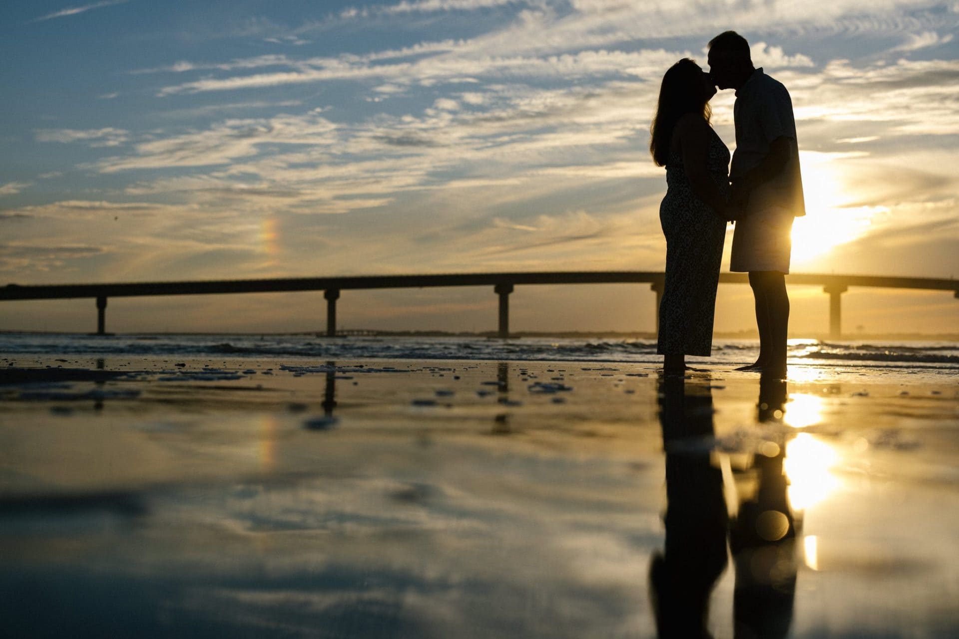 A silhouette of a mom and dad kissing on the wet sand with the low sun coming through between them and the bridge running across behind in Ocean City, New Jersey