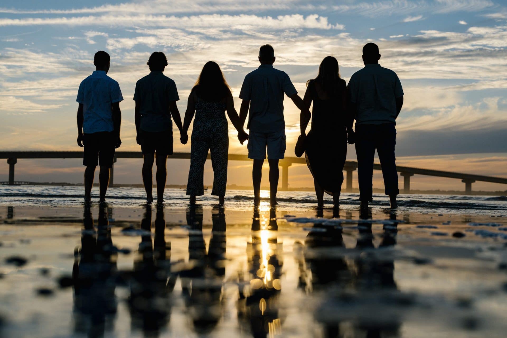 A row of family members in silhouette on the wet sand, backs to the camera, facing the low light with the bridge running off behind them in Ocean City, New Jersey