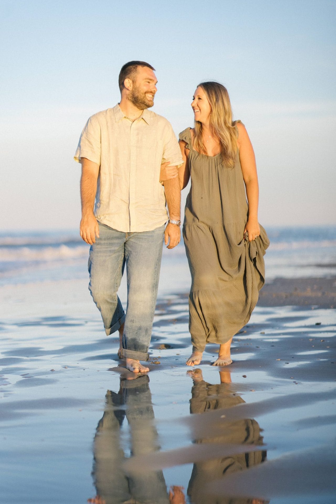 A couple walking barefoot along the water line, looking at each other and laughing, with their reflection on the wet sand in Ocean City, New Jersey