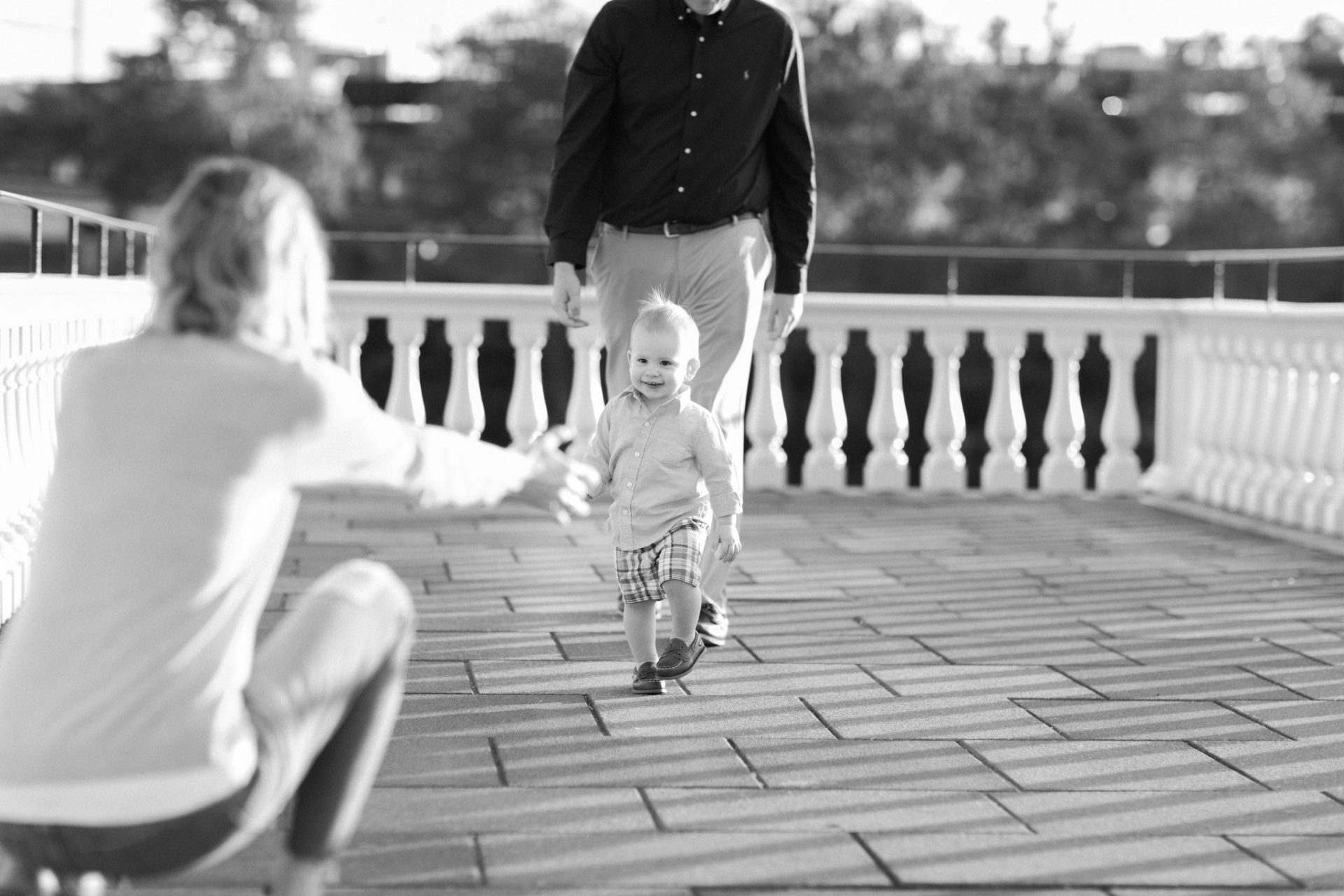 A black and white photograph of a small boy toddling toward his mom, who is crouched down with one arm out, as his dad walks behind him in Philadelphia