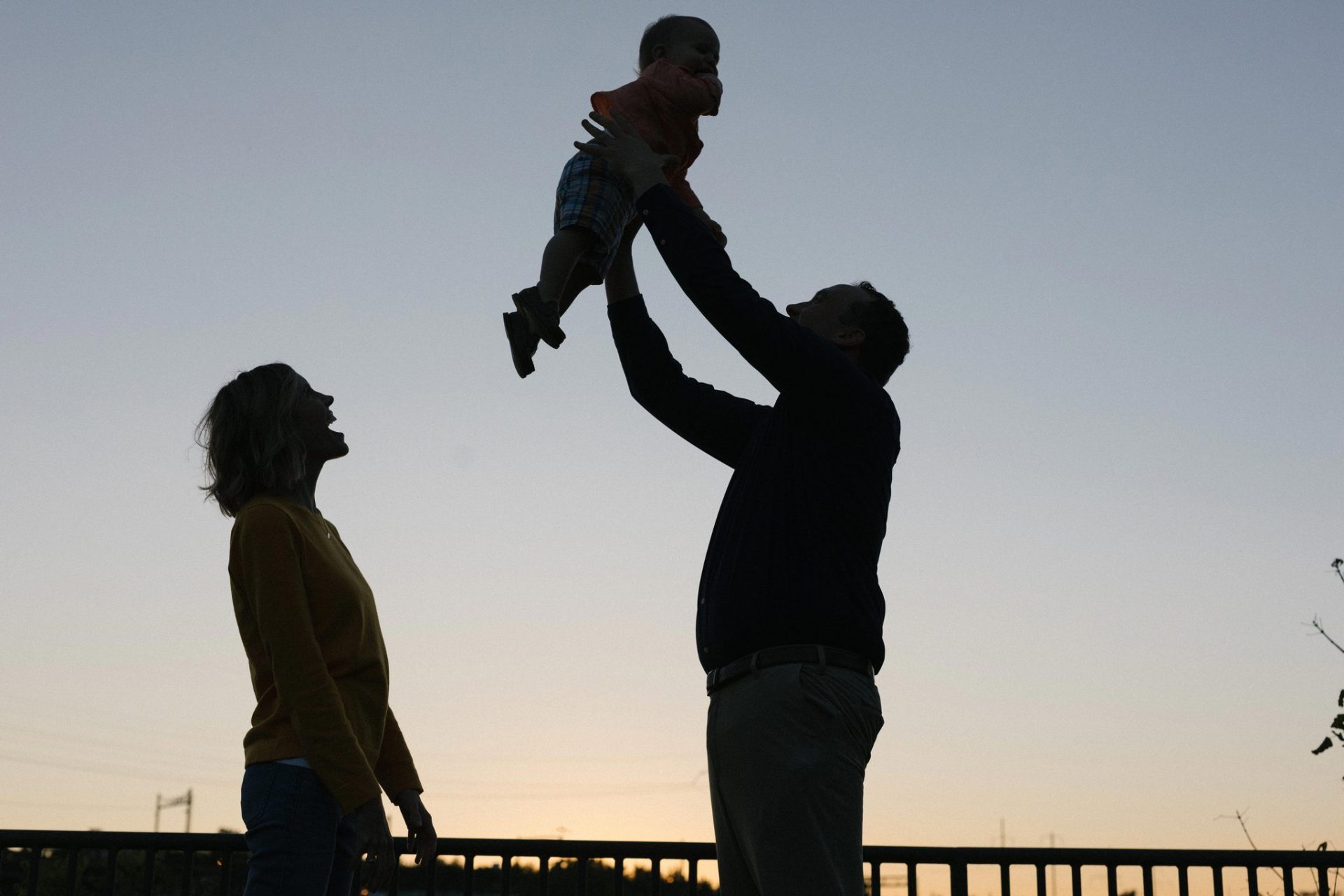 A backlit photograph of a dad lifting his son into the air at sunset while his wife laughs beside them in Philadelphia